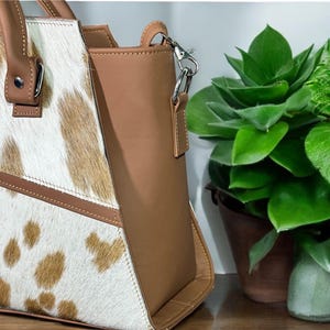 May include: A brown and white cowhide tote bag with a brown leather trim and a silver chain strap.