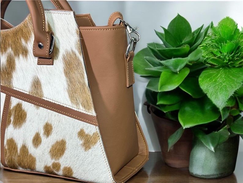May include: A brown and white cowhide tote bag with a brown leather trim and a silver chain strap.