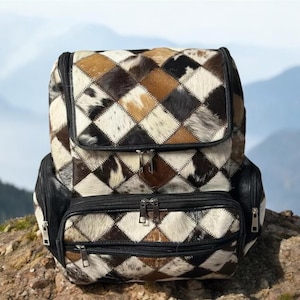 May include: A black leather backpack with a patchwork design of brown, white, and black cowhide. The backpack has multiple pockets and zippers.