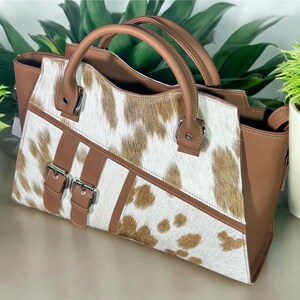 May include: A brown and white cowhide handbag with two buckles and a top handle.