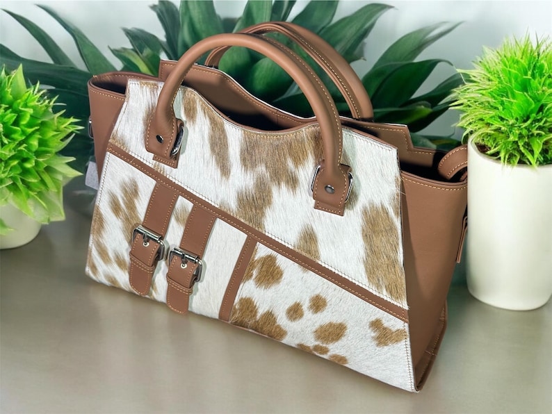 May include: A brown and white cowhide handbag with two buckles and a top handle.