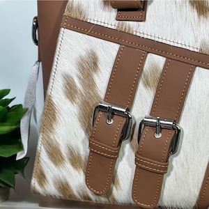 May include: Close-up of a brown leather handbag with a white and brown cowhide pattern. The bag has two silver buckles and brown straps.