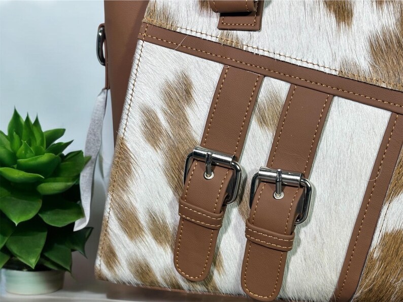 May include: Close-up of a brown leather handbag with a white and brown cowhide pattern. The bag has two silver buckles and brown straps.
