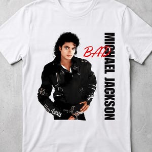 May include: A white short-sleeved t-shirt featuring a graphic of Michael Jackson wearing a black leather jacket with buckles and zippers. The word BAD is printed in red script, and the name MICHAEL JACKSON is printed vertically in black text.