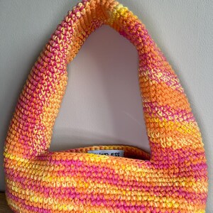 May include: A colourful crochet bag in shades of orange, yellow, and pink. The bag has a rounded handle and a rectangular body. The bag has a label that reads "less".