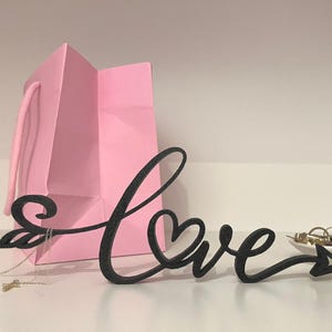 Könnte beinhalten: Ein schwarzes, glitzerndes "Love"-Schild mit Pfeildesign, auf einer weißen Oberfläche. Im Hintergrund befindet sich eine rosa Geschenktüte sowie eine kleine Schale mit Schmuck. Das Schild ist ein dekorativer Gegenstand.