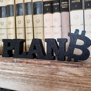 Könnte beinhalten: Schwarze, strukturierte Buchstaben mit der Aufschrift "PLAN B" mit dem Bitcoin-Symbol. Die Buchstaben stehen auf einem Holzregal vor einer Reihe alter Bücher. Das Bitcoin-Symbol ist der letzte Buchstabe.