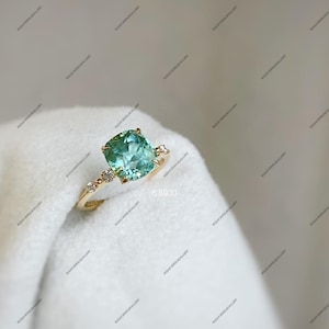 May include: A gold ring featuring a large, square-cut, light teal gemstone. The ring has a delicate band with small accent stones and the text "8 Bijou".