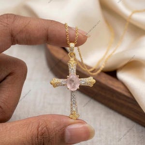 Könnte beinhalten: Eine goldfarbene Kreuz-Anhänger-Halskette mit einem Rosenquarz-Mittelstein und kleinen, klaren Steinen. Das Kreuz hat aufwendige Details und hängt an einer zarten Goldkette. Der Schmuck wird vor einem weichen, neutralen Hintergrund präsentiert.