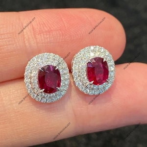 Può includere: Un paio di orecchini a lobo con rubini. Ogni orecchino presenta un rubino rosso ovale al centro, circondato da un doppio alone di piccoli diamanti scintillanti. Gli orecchini sono incastonati in un metallo bianco, probabilmente platino o oro bianco.