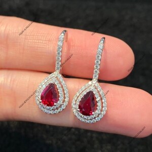 May include: A pair of elegant drop earrings featuring pear-shaped, deep red gemstones surrounded by a halo of smaller, clear stones. The earrings have a slender, curved top section also adorned with clear stones, creating a sparkling, luxurious look.