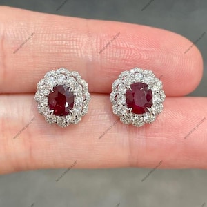 May include: A pair of stud earrings featuring a central, deep red gemstone surrounded by a halo of smaller, clear gemstones. The earrings have a classic, elegant design, perfect for adding a touch of sparkle.