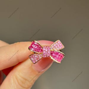 May include: A gold-toned bow ring with pink gemstones. The bow design features a central cluster of smaller stones and larger, square-cut stones forming the bow's loops. The ring is held by a hand. The text "ROZANJEWELSGALLERY" is visible.