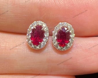 Könnte beinhalten: Ein Paar ovale Rubinohrringe. Jeder Ohrring hat einen leuchtend roten Rubin-Mittelstein, der von einem Halo aus kleinen, funkelnden weißen Diamanten umgeben ist. Die Ohrringe sind in einem hellen Metall gefasst.