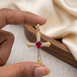 May include: Gold-tone cross pendant with a central oval ruby and small clear stones. The cross has ornate detailing and is held between fingers against a soft, neutral background. A religious jewelry item.