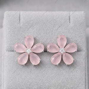 Puede incluir: Un par de pendientes con forma de flor. Cada pendiente presenta cinco pétalos de color rosa claro que rodean un acento central brillante. Los pendientes están engarzados en un soporte de metal plateado, presentados sobre una superficie gris texturizada.
