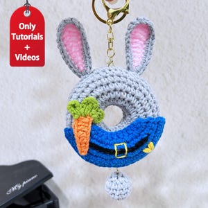 May include: A handmade crochet bunny donut keychain with gray, pink, orange, green, and blue accents. The keychain features a carrot, a blue hat with a gold buckle, and a small gray ball. The tag reads "Only Tutorials + Videos".
