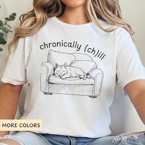 May include: White t-shirt featuring a black and white illustration of a cat napping in an armchair. The text "chronically (ch)ill" is printed on the shirt.