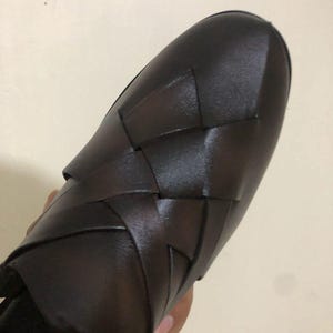 May include: A dark brown slip-on shoe featuring a woven leather upper with wide, interlocking straps that create a textured, geometric pattern across the top. The shoe has a rounded toe and a smooth, polished finish on the leather surface.