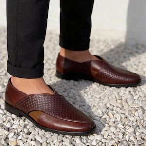 Pu&ograve; includere: Un paio di scarpe slip-on in pelle marrone scuro con una trama intrecciata sui pannelli superiori, una punta liscia in pelle e un tacco basso nero. Le scarpe presentano un caratteristico design asimmetrico ritagliato sui lati e una punta tonda.
