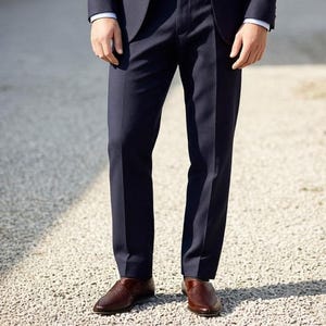 Pu&ograve; includere: Un paio di pantaloni eleganti blu navy scuro dal taglio dritto, abbinati a scarpe slip-on in pelle intrecciata marrone. I pantaloni presentano una piega stirata al centro di ogni gamba e le scarpe hanno una punta arrotondata.