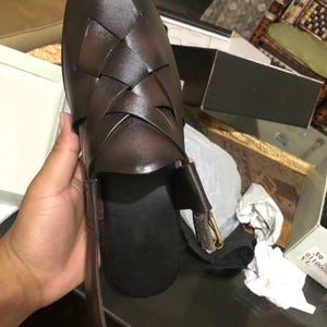 May include: A dark brown woven leather sandal featuring a closed toe with an intricate lattice pattern, a black footbed, and an adjustable ankle strap with a metallic buckle. The shoe is displayed with a visible size 39 label on the packaging box.