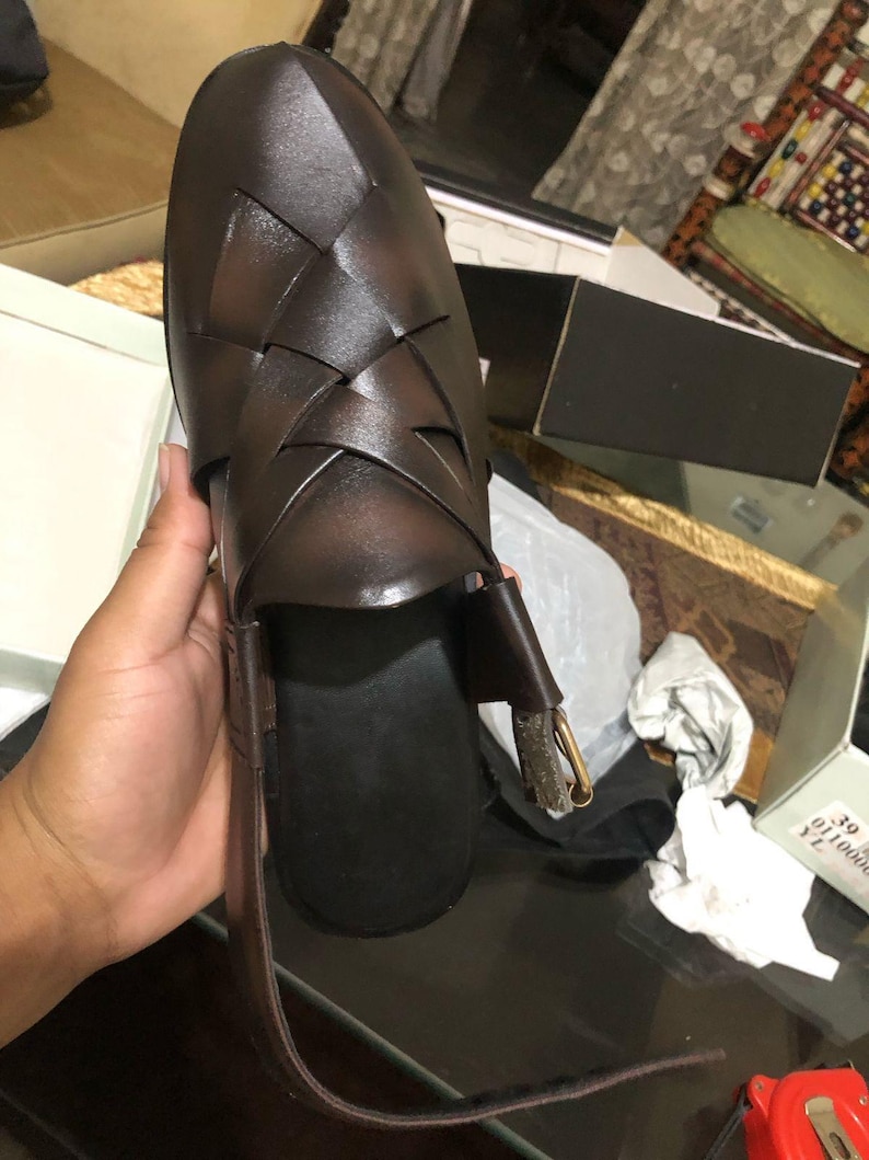 May include: A dark brown woven leather sandal featuring a closed toe with an intricate lattice pattern, a black footbed, and an adjustable ankle strap with a metallic buckle. The shoe is displayed with a visible size 39 label on the packaging box.