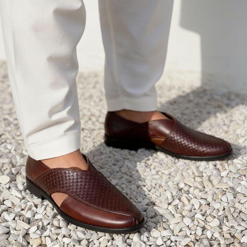 Pu&ograve; includere: Un paio di mocassini in pelle marrone scuro con una trama intrecciata sulla parte superiore, punta affusolata e aperture laterali. Le scarpe sono indossate con pantaloni bianchi, caratterizzate da un design elegante con tacco basso e liscio.
