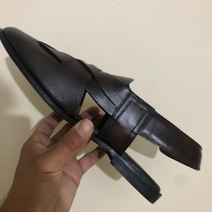 May include: A dark brown leather sandal featuring a closed-toe design with wide, interwoven straps across the top, a flat sole, and a secure heel strap. The sandal has a smooth finish and a structured silhouette, showcasing a classic open-side construction.