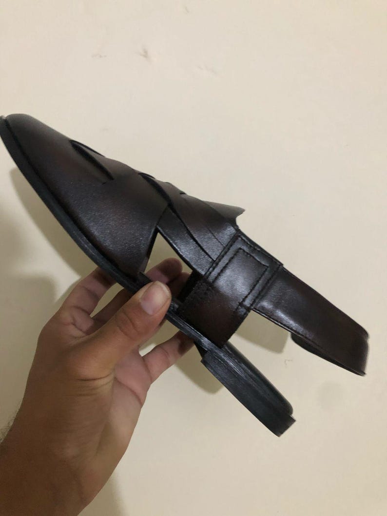 May include: A dark brown leather sandal featuring a closed-toe design with wide, interwoven straps across the top, a flat sole, and a secure heel strap. The sandal has a smooth finish and a structured silhouette, showcasing a classic open-side construction.