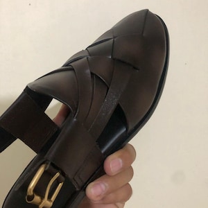 May include: A dark brown woven leather sandal featuring a closed-toe design with wide interlaced straps. The sandal includes a side strap with a gold-toned metal buckle closure and a smooth, dark brown footbed for a secure and adjustable fit.