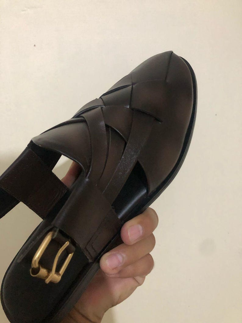 May include: A dark brown woven leather sandal featuring a closed-toe design with wide interlaced straps. The sandal includes a side strap with a gold-toned metal buckle closure and a smooth, dark brown footbed for a secure and adjustable fit.