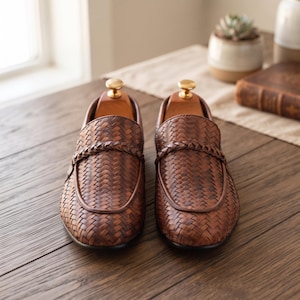 May include: A pair of brown leather loafers featuring a woven texture across the upper surface, a braided leather strap detail across the vamp, and a wooden shoe tree inserted into each shoe to maintain the shape of the footwear.