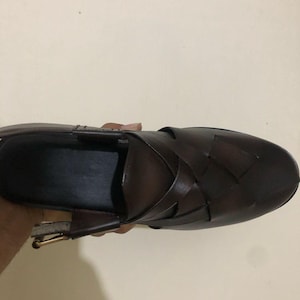 May include: A dark brown leather shoe featuring a woven upper design with wide, interlaced straps. The shoe has a closed, rounded toe, an open heel area, and a metallic buckle closure on the side strap, showcasing a smooth, polished finish.