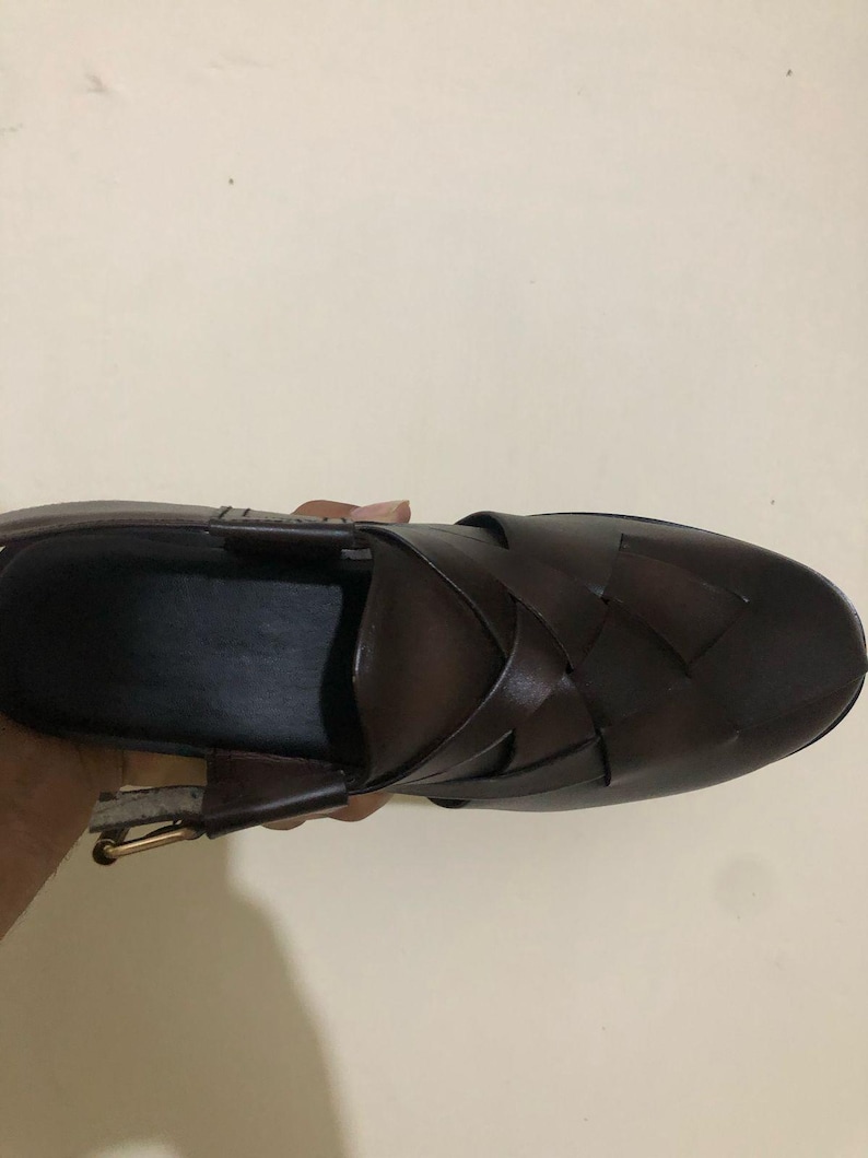 May include: A dark brown leather shoe featuring a woven upper design with wide, interlaced straps. The shoe has a closed, rounded toe, an open heel area, and a metallic buckle closure on the side strap, showcasing a smooth, polished finish.