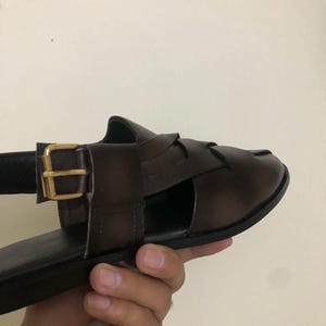 May include: A dark brown leather sandal featuring a woven strap design across the top and a side ankle strap secured with a gold-toned metal buckle. The sandal has a flat sole and a smooth, polished finish on the leather upper material.