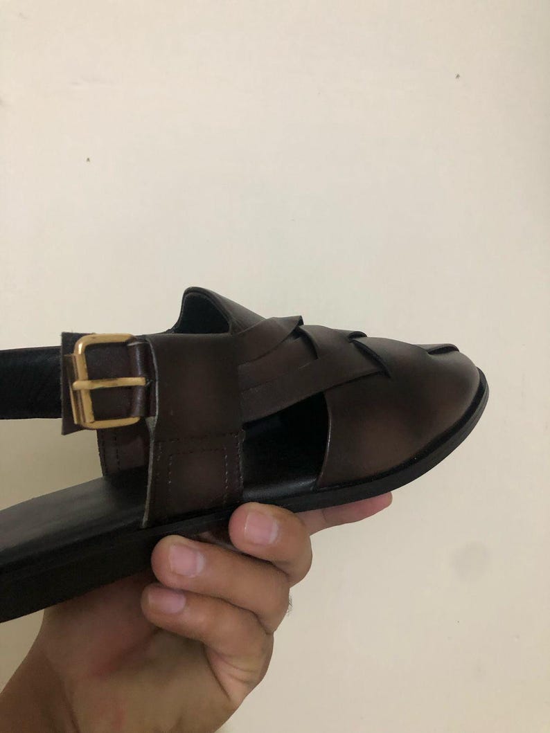 May include: A dark brown leather sandal featuring a woven strap design across the top and a side ankle strap secured with a gold-toned metal buckle. The sandal has a flat sole and a smooth, polished finish on the leather upper material.