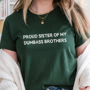 May include: Dark green t-shirt with the text "PROUD SISTER OF MY DUMBASS BROTHERS" in white. The shirt is a crew neck style and is made of a soft material. The shirt is being worn by a person.