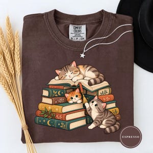 May include: A dark brown crew neck t-shirt featuring a graphic of three cats resting on a stack of colorful books. One cat sleeps on top, one peeks from between the books, and one sits beside the stack. The shirt has a Comfort Colors brand label.