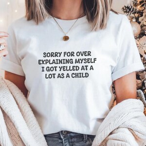 May include: White t-shirt with the text "SORRY FOR OVER EXPLAINING MYSELF I GOT YELLED AT A LOT AS A CHILD" in black. The shirt is worn by a person, with a gold necklace and a light-colored sweater draped over the shoulders.