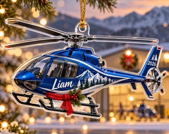 May include: A blue helicopter Christmas ornament featuring the name Liam in white script, a mountain and pine tree graphic, and the year 2026 on the tail. The helicopter is decorated with a small red knit scarf and green holly sprigs.