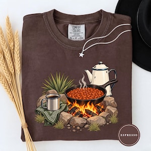 May include: A brown crewneck t-shirt featuring a graphic of a campfire with a cast iron skillet of beans, a white coffee pot, and a metal mug on a green bandana. The shirt has a Comfort Colors label at the neck and a silver star pendant necklace.
