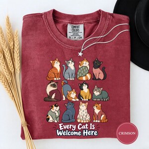 May include: A crimson crew neck t-shirt featuring a graphic of twelve illustrated cats in various poses and patterns. Below the cats, the text reads "Every Cat Is Welcome Here". The shirt has a Comfort Colors label inside the collar.