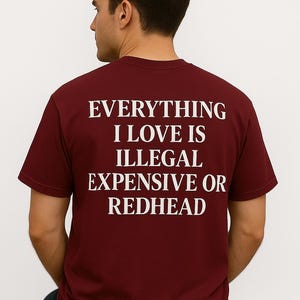 May include: Burgundy t-shirt with white text that reads "EVERYTHING I LOVE IS ILLEGAL EXPENSIVE OR REDHEAD." The shirt is a crew neck and is being worn by a person.