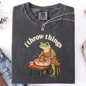 May include: A charcoal gray t-shirt featuring a graphic of a green frog wearing a red and white striped apron, sitting on a stool and working at a pottery wheel. The text "i throw things" is printed in a cream-colored font above the frog illustration.