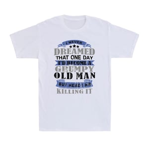 May include: White t-shirt with a humorous graphic. The design features the text "I NEVER DREAMED THAT ONE DAY I'D BECOME A GRUMPY OLD MAN BUT HERE I AM KILLING IT" in gray, blue, and black.