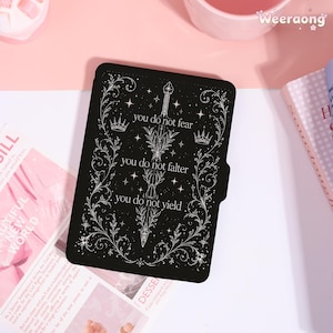 May include: A black e-reader case featuring a white line art illustration of a sword surrounded by ornate floral vines, stars, and two crowns. The text reads: you do not fear, you do not falter, you do not yield. The case has a rectangular shape.