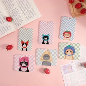 Può includere: Una collezione di custodie per telefoni con gatti dei cartoni animati che indossano cappelli. Ogni custodia ha uno sfondo a scacchi in vari colori, con i gatti raffigurati in diversi abiti, tra cui un pomodoro, un orso e un cappello di formaggio.