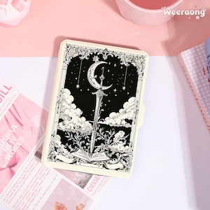 May include: A rectangular e-reader case featuring a black and white illustration of a sword piercing an open book beneath a crescent moon and stars. The design is framed by ornate scrollwork and clouds, with a cream-colored border and edges.