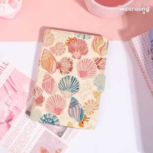 May include: A beige notebook cover adorned with colorful seashell illustrations in shades of pink, blue, yellow, and red. The notebook is open, revealing pages and text from a magazine. The cover has a magnetic closure.